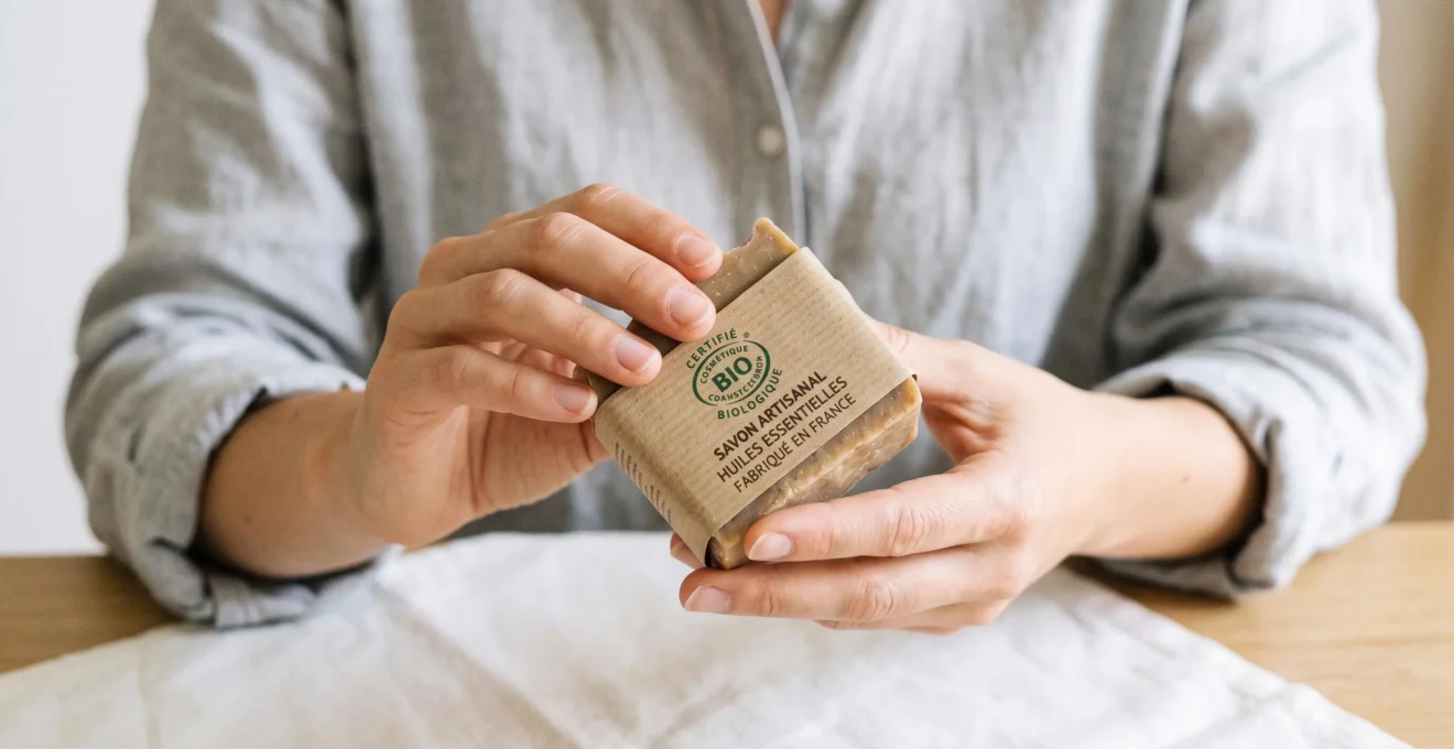 Mains d'une femme tenant un savon solide emballé dans du papier kraft avec logo de certification bio Ecocert ou Cosmébio clairement visible